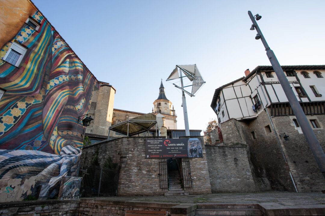 Fotografía del casco antiguo de Vitoria-Gasteiz, con un mural colorido en la fachada de un edificio, la torre de la iglesia de Santa María al fondo y un cartel de entrada a la catedral.