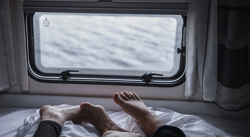 Dos personas recostadas en una cama dentro de una furgoneta, con vista hacia fuera a través de una ventana rectangular.